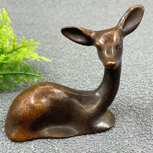 Könnte beinhalten: Bronze-farbene Hirschfigur in Ruheposition. Die Skulptur hat einen schlanken Hals, große Ohren und einen detaillierten Körper. Der Hirsch sitzt auf einer strukturierten Oberfläche, mit einer grünen Pflanze im Hintergrund.