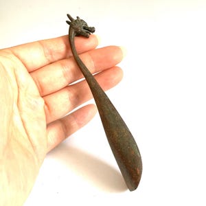Pode incluir: Uma colher de cor bronze com cabeça de dragão e cabo longo e curvo. A cabeça da colher apresenta detalhes de dragão e o cabo afunila para uma extremidade arredondada. A colher é segurada contra um fundo branco.