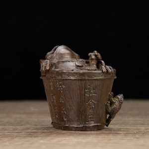 Puede incluir: Una escultura en forma de cubo de color bronce con detalles intrincados. El cubo tiene una tapa y una peque&ntilde;a criatura trepando por un lado. La superficie est&aacute; texturizada con caracteres chinos.