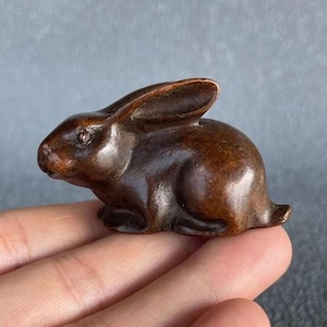 Op de afbeelding: Een kleine, bruine konijnenfiguur. De sculptuur is gedetailleerd, met zichtbare oren, ogen en poten. Het konijn zit, met de oren omhoog. Het beeldje heeft een gladde, gepolijste afwerking en lijkt van hout of een vergelijkbaar materiaal te zijn.