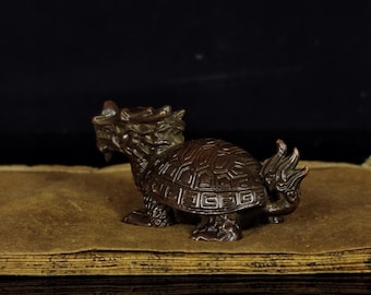 May include: A bronze-colored figurine of a turtle with a dragon-like head and tail. The turtle has a detailed shell pattern and is sitting on a textured, brown surface. The figurine is a decorative item.