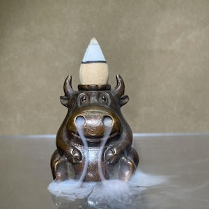 May include: A bronze bull-shaped incense burner with smoke billowing from the nostrils. The incense cone is beige with a blue and white top. The burner has a detailed design and is on a reflective surface.
