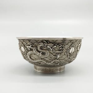 May include: A silver-colored bowl with intricate dragon and wave pattern relief. The bowl has a wide rim and a pedestal base with a geometric design. The dragon is depicted in a detailed, three-dimensional style, adding depth to the design.