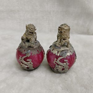 May include: Two antique, decorative bottles with a red, spherical body and silver-colored metal accents. Each bottle features a detailed, small lion figure on top and a dragon design around the base. The bottles are displayed on a light-colored surface.