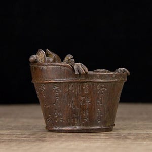 Puede incluir: Un peque&ntilde;o cubo de madera de estilo antiguo con detalles tallados y una peque&ntilde;a criatura esculpida en el borde. El cubo tiene texto grabado alrededor del exterior y se encuentra sobre una superficie de madera.