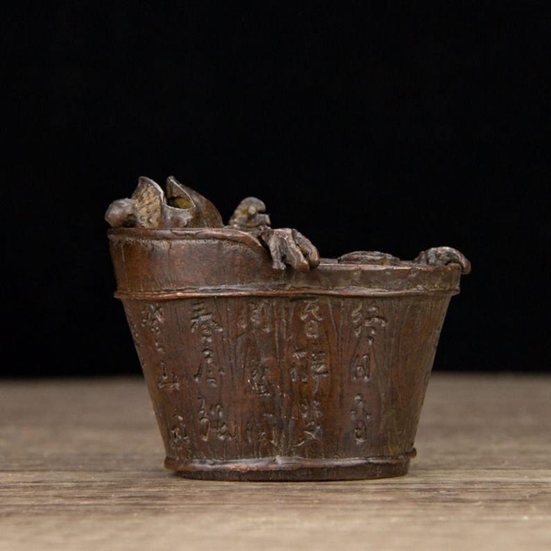 Puede incluir: Un peque&ntilde;o cubo de madera de estilo antiguo con detalles tallados y una peque&ntilde;a criatura esculpida en el borde. El cubo tiene texto grabado alrededor del exterior y se encuentra sobre una superficie de madera.