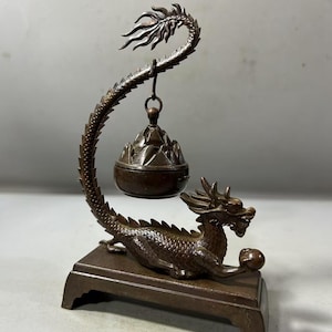 Puede incluir: Quemador de incienso de dragón de bronce con un cuerpo curvo y detallado y un porta incienso colgante. El dragón descansa sobre una base rectangular, sosteniendo una esfera. El porta incienso está suspendido de la cola del dragón.