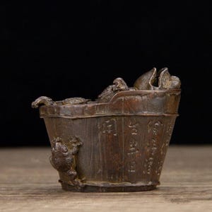 Puede incluir: Un cubo en miniatura de color bronce con una rana trepando por el lateral. El cubo tiene una textura de madera y est&aacute; lleno de elementos esculpidos, posiblemente representando una escena. Hay caracteres chinos grabados en el lateral.