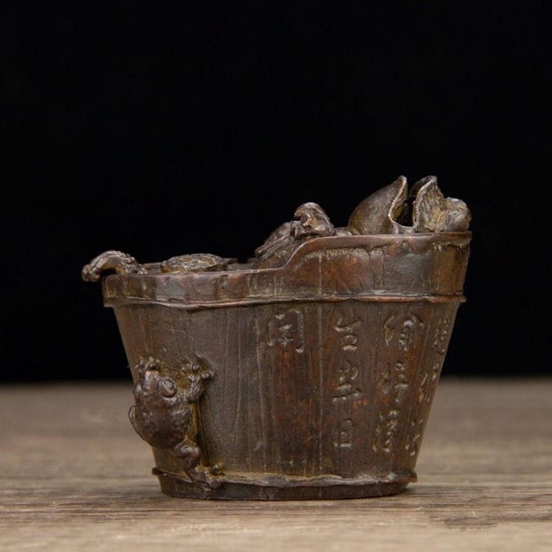 Puede incluir: Un cubo en miniatura de color bronce con una rana trepando por el lateral. El cubo tiene una textura de madera y est&aacute; lleno de elementos esculpidos, posiblemente representando una escena. Hay caracteres chinos grabados en el lateral.