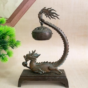 Puede incluir: Quemador de incienso de dragón de bronce. El dragón está sobre una base rectangular, con un cuerpo curvado que sostiene un cuenco con tapa. El dragón sostiene una esfera. El quemador de incienso es un objeto decorativo.