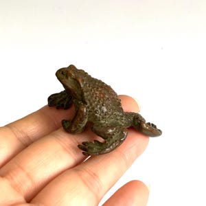 Könnte beinhalten: Kleine, bronzefarbene Froschfigur mit strukturierter Oberfläche. Der Frosch befindet sich in einer Ruheposition, die Beine sind ausgestreckt. Die Figur ist wahrscheinlich aus Metall und hat ein detailliertes, realistisches Aussehen.