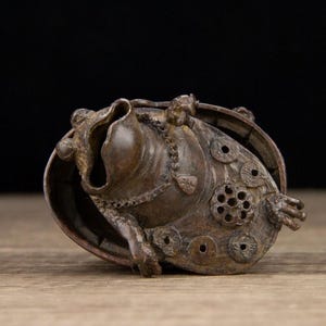 Puede incluir: Escultura de bronce de una criatura con forma de rana, con detalles intrincados. La escultura presenta una gran boca abierta, detalles similares a monedas y una superficie texturizada. El dise&ntilde;o sugiere un objeto decorativo o simb&oacute;lico.