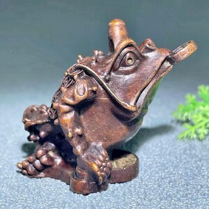 Könnte beinhalten: Detaillierte, braune Holzfrosch-Skulptur mit einer Münze im Maul. Der Frosch hat strukturierte Haut, detaillierte Augen und sitzt auf einem kleinen, runden Sockel. Die Skulptur ist ein dekoratives Objekt, möglicherweise für einen Schreibtisch oder ein Regal.