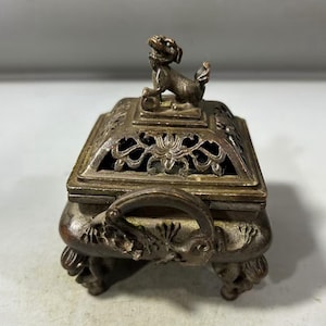 May include: A bronze-colored incense burner with a decorative, openwork design. The square-shaped burner features a small dog figurine on top and ornate handles. The burner rests on four decorative feet, showcasing an antique aesthetic.
