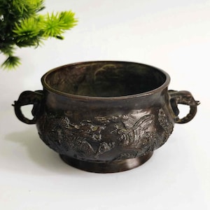 Pode incluir: Queimador de incenso redondo, cor bronze, com alças em forma de elefante. O exterior apresenta entalhes em relevo detalhados de dragões e fénix. O vaso assenta numa pequena base circular.