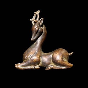Puede incluir: Figura de ciervo de bronce en posición de descanso, con cuernos detallados y cuello esbelto. La escultura tiene un aspecto envejecido y antiguo, con textura visible y pátina marrón oscuro. El ciervo está tumbado, con las patas recogidas.