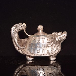 May include: A silver-colored, turtle-shaped decorative object with dragon heads at each end. The turtle's shell is inscribed with Chinese characters. The object has a lid with a small handle and is set against a black background.