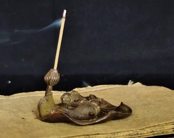 May include: A bronze incense holder featuring a fish design on a leaf-shaped base. An incense stick is lit, emitting a wisp of smoke. The holder sits on a stack of aged, yellowed pages, creating a vintage aesthetic.