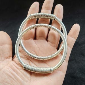 May include: Two silver bangles with engraved Chinese characters. The bangles are round and have a smooth, polished finish. The text is etched around the circumference of the bangles. The bangles are displayed on a hand.