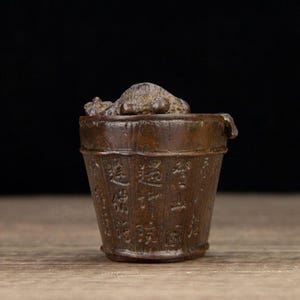 Puede incluir: Un peque&ntilde;o cubo de madera de estilo antiguo con caracteres chinos tallados. El cubo es de color marr&oacute;n oscuro y tiene una tapa con una peque&ntilde;a escultura texturizada en la parte superior. El cubo probablemente se utiliza para almacenamiento o fines decorativos.