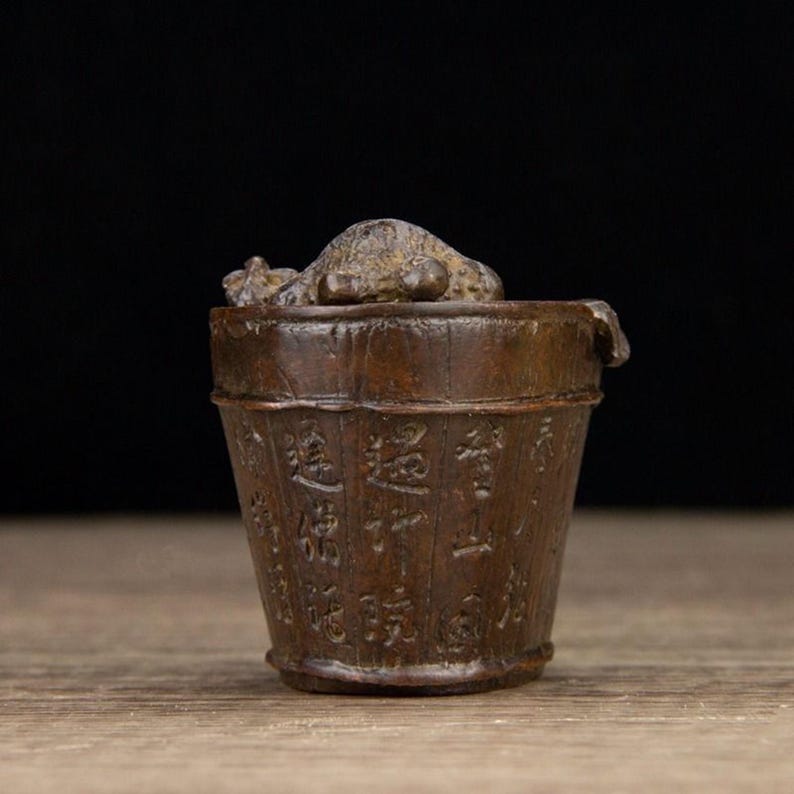 Puede incluir: Un peque&ntilde;o cubo de madera de estilo antiguo con caracteres chinos tallados. El cubo es de color marr&oacute;n oscuro y tiene una tapa con una peque&ntilde;a escultura texturizada en la parte superior. El cubo probablemente se utiliza para almacenamiento o fines decorativos.