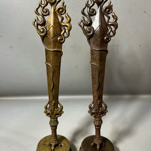 May include: Two antique bronze ritual objects. Each has a flame-like top, a long, tapered body, and a decorative base. One is gold-toned, the other is bronze. The objects are likely used for religious or ceremonial purposes.