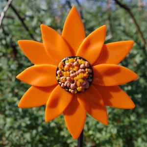 May include: A bright orange glass flower with a center made of small, colorful glass pieces. The flower has 12 petals and is on a black metal stem.