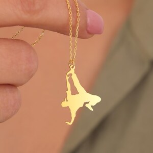 Könnte beinhalten: Eine goldfarbene Halskette mit einem Anhänger, der die Silhouette eines Breakdancers zeigt. Der Anhänger hängt an einer filigranen Kette. Der Breakdancer ist in einer dynamischen Pose dargestellt, mit einem ausgestreckten Arm und sich bewegenden Beinen.
