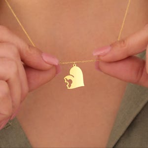 May include: A gold-colored necklace featuring a heart-shaped pendant with a cut-out design. The pendant hangs from a delicate chain. The necklace is being held up by a person.