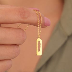 May include: A gold-colored necklace with a rectangular pendant featuring a feather cutout. The delicate chain and pendant are held up by a hand with painted nails. The necklace is a piece of jewelry.