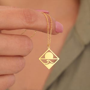 Könnte beinhalten: Goldfarbene Halskette mit einem Anhänger in Rautenform. Der Anhänger zeigt ein Sonnen- und Wellenmotiv. Die Kette ist filigran. Die Halskette wird von den Fingern einer Person gehalten.