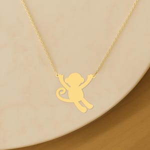 May include: A gold-colored necklace featuring a monkey pendant. The monkey is depicted in a playful pose with its arms outstretched and a curled tail. The necklace chain is delicate, and the pendant is smooth and shiny.