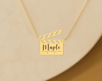 May include: A gold-colored necklace featuring a film clapperboard pendant. The clapperboard has the name "Maple" engraved on it. The necklace is on a gold chain, and the background is a light beige color.