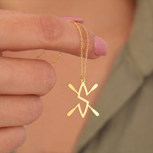 May include: A gold-colored necklace with a pendant in the shape of a stylized star or geometric design. The necklace is held by a person's fingers, showcasing the delicate chain and pendant's intricate details. The background is blurred, focusing attention on the jewelry.