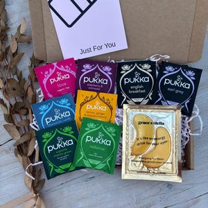 Könnte beinhalten: Eine Geschenkbox mit Pukka Teebeuteln in verschiedenen Geschmacksrichtungen, darunter Love, English Breakfast und Radiance. Ebenfalls enthalten sind Augenmasken von Grace & Stella. Auf der Box steht "Just For You".