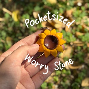 Könnte beinhalten: Ein sonnenblumenförmiger Sorgenstein in Gelb und Braun. Der Stein wird in einer Hand vor einem verschwommenen grün-gelben Hintergrund gehalten. Der Text "Pocketsized Worry Stone" ist auf dem Bild geschrieben.