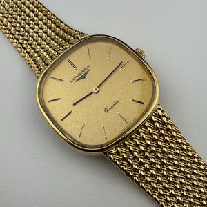 Könnte beinhalten: Eine goldfarbene Longines Quartz-Uhr mit quadratischem Zifferblatt und Mesh-Armband. Das Zifferblatt zeigt das Longines-Logo und das Wort "Quartz". Das Armband ist in einem gewebten Design gehalten, und die Uhr hat einen klassischen, eleganten Stil.