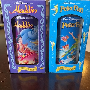 May include: Two collector's series Disney glasses in their original boxes. One box is purple with "Aladdin" in gold lettering, and the other is blue with "Peter Pan" in gold lettering. The glasses feature colorful illustrations from the classic Disney films.