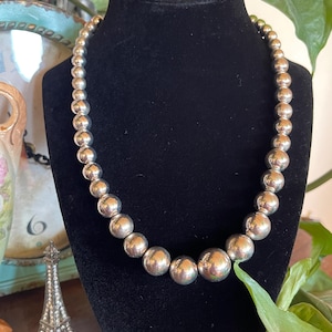 May include: A graduated silver-tone bead necklace featuring polished spherical beads that increase in size from the clasp area toward the center, displayed on a black velvet-covered neck form for clear visibility of the metallic finish.