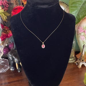 May include: A gold chain necklace featuring an oval-shaped red gemstone pendant surrounded by a halo of small white stones. The pendant hangs from a delicate gold chain, displayed on a black velvet-textured bust form.