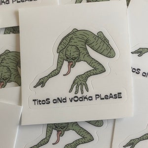 May include: A sticker with a green, striped, humanoid creature with a long tongue crawling on all fours. The text "Titos and Vodka Please" is printed on the sticker.
