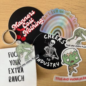 May include: A selection of stickers and a keyring. Stickers feature text like "Manners Cost Nothing", "don't tell me what to do" on a rainbow, a skeleton with "Cheers to Industry", and a zombie. A keyring with a green creature is also visible.