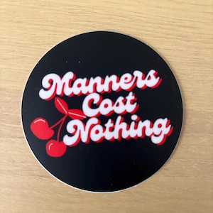 May include: A round sticker with the phrase "Manners Cost Nothing" in white, outlined in red, on a black background. A pair of red cherries with green stems are featured below the text. The sticker is on a light brown wooden surface.