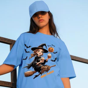 Op de afbeelding: Lichtblauw T-shirt met een Halloween-thema. Het ontwerp toont een cartoon heks op een bezemsteel met koffie, met vleermuizen en een pompoen. De tekst "Brew, Please" staat boven de pompoen en onder de heks.