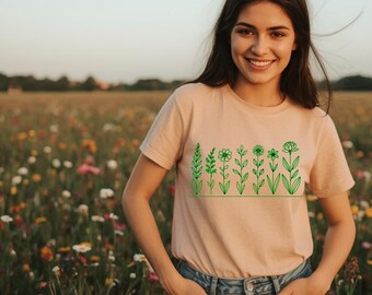 May include: A light peach-colored t-shirt featuring a green line drawing of various flowers and plants. The design is centered on the shirt. The person is wearing blue jeans and is smiling in a field of wildflowers.