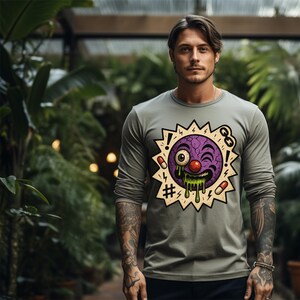 Puede incluir: Una camiseta de manga larga verde oliva con un dibujo animado. El dibujo animado es una cara morada con una nariz roja, un ojo y baba verde, rodeada por una estrella amarilla con signos de exclamación, pastillas y un símbolo de hashtag.