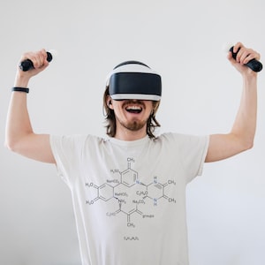 Puede incluir: Una persona con un casco de realidad virtual y controladores, con una camiseta de color claro con un diagrama de estructura química. La persona está sonriendo y parece estar disfrutando de una experiencia de realidad virtual.