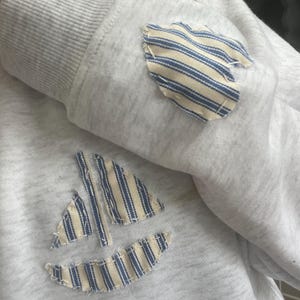 May include: A pale grey sweatshirt featuring a sailboat applique and a small patch. The sailboat and patch are made of fabric with blue and white stripes. The sweatshirt has a ribbed cuff.