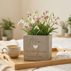May include: A square, gray concrete planter with the words "Mom", "LOVE GROWS HERE", and "caroline" on the front, holding white and pink flowers. A wooden tray holds the planter, a mug, and a Mother's Day card.