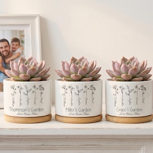 Peut inclure: Trois pots de plantes succulentes en céramique blanche avec des bases en bois, chacun avec un nom différent : Thompson's Garden, Miller's Garden et Grace's Garden. Les pots ont un motif floral et le texte "Love Grows Here". Chaque pot contient une plante succulente violet clair.